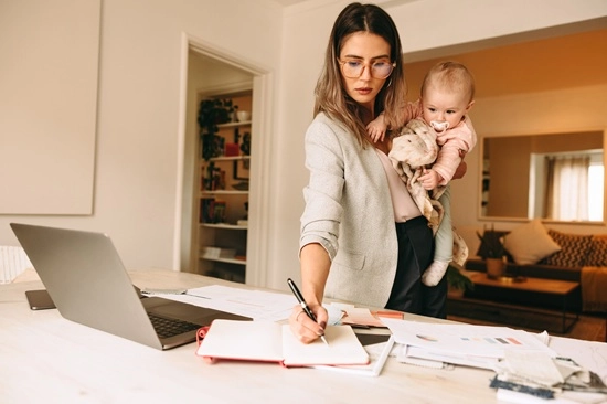 Mother holding her baby while working at a laptop and notes