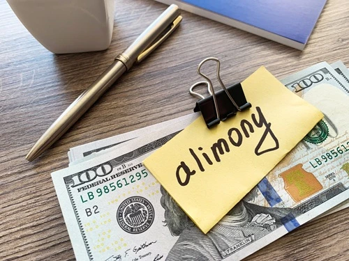 Note reading alimony clipped to hundred-dollar bills beside a pen