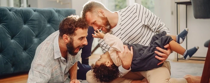 Two fathers playing with their laughing toddler on a sofa