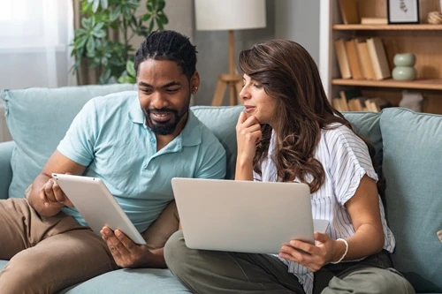 Couple on a sofa discussing paperwork on a laptop and tablet