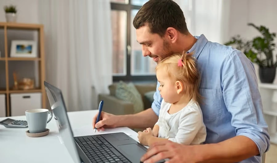 Father signing papers at a laptop with his daughter on his lap